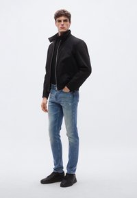 Čierna bomber bunda so zapínaním na zips a bočnými vreckami, v kombinácii s svetlomodrými džínsami s opotrebovaným vzhľadom a čiernymi teniskami. Jednoduché pozadie.