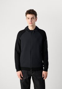 Črna zip-up kapuca z teksturirano sprednjo stranjo in gladkimi rokavi. Ima kapuco, vrvici in žep na levi roki.