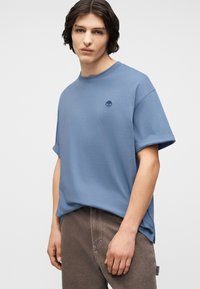 Νέος άνδρας που φοράει ανοιχτό μπλε φαρδιά μπλουζάκι με μικρό λογότυπο στο στήθος και καφέ παντελόνι, στέκεται μπροστά σε απλό φόντο.