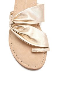 Sandalia plana metálica dorada con parte superior de tela anudada y suela de cuero beige. Presenta un diseño minimalista y detalles de costura.
