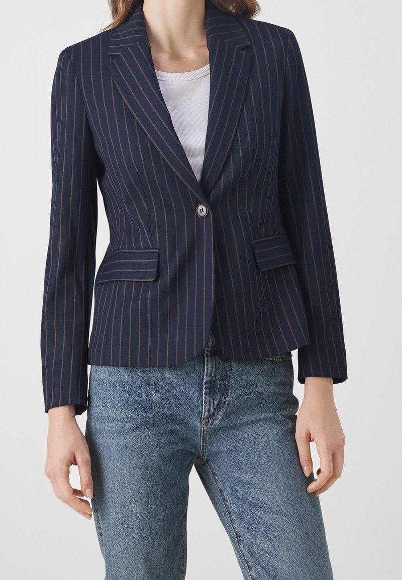 Femme portant un blazer bleu marine à fines rayures avec un bouton, par-dessus un haut blanc et un jean bleu, sur un fond uni.