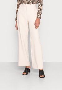 Béžové kalhoty s širokými nohavicemi a vysokým pasem, vyrobené z hladkého materiálu. V kombinaci s černými sandály na podpatku s otevřenou špičkou. Viditelný květinový top s dlouhým rukávem.