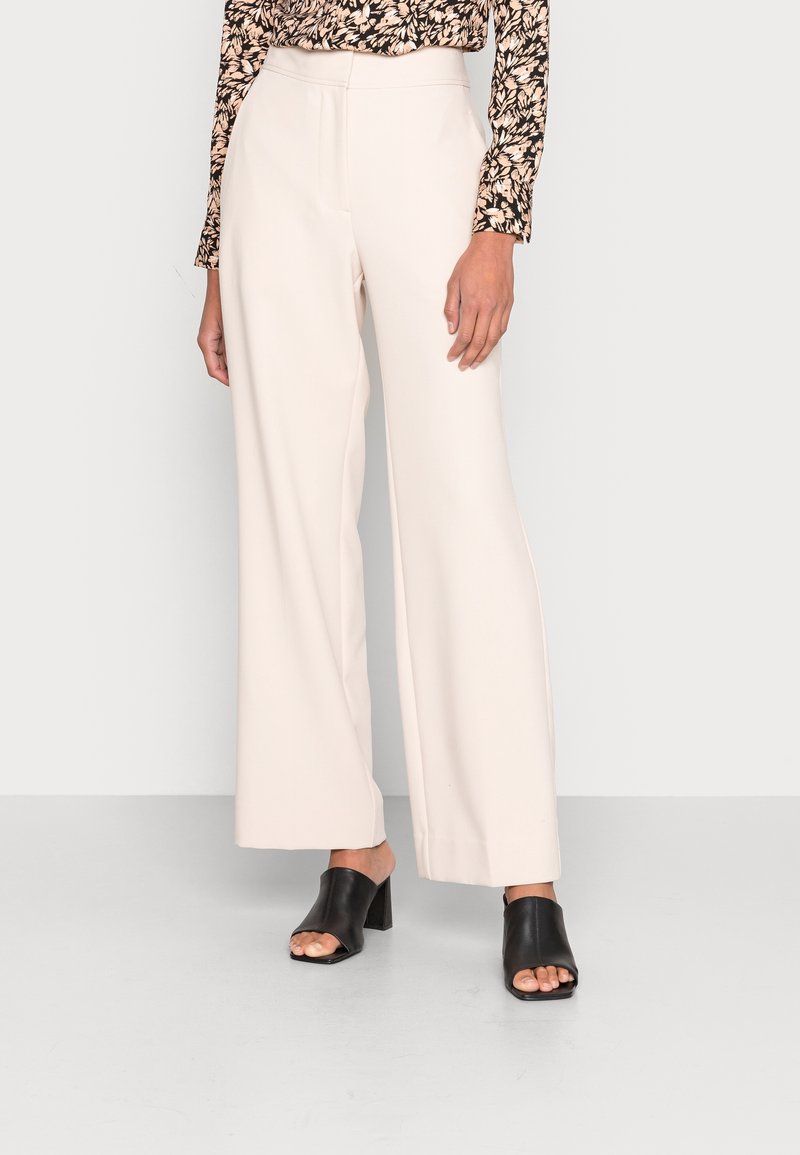 Béžové kalhoty s širokými nohavicemi a vysokým pasem, vyrobené z hladkého materiálu. V kombinaci s černými sandály na podpatku s otevřenou špičkou. Viditelný květinový top s dlouhým rukávem.