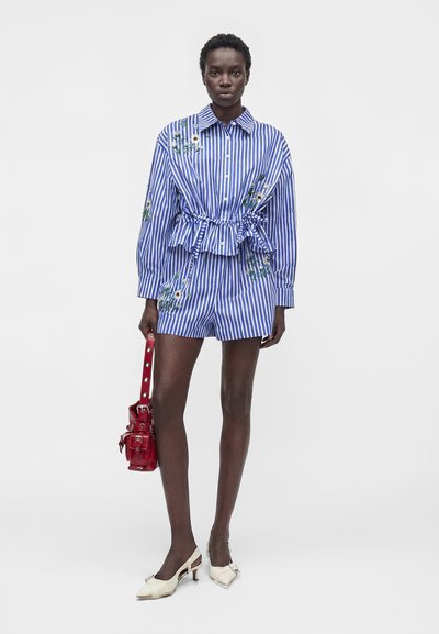 Femme grande portant un haut rayé bleu et blanc brodé de fleurs et un short assorti, tenant un sac à main rouge et portant des chaussures blanches à talons.