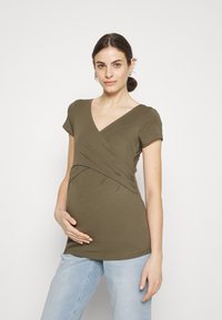 Olivenfarget gravidebluse med korte ermer, v-halsdesign og rynket stoff rundt midjen, sammen med lyseblå jeans.