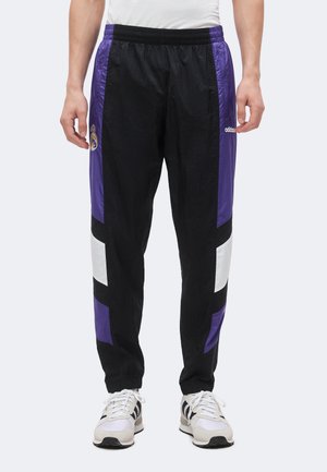 Personne portant un pantalon de survêtement noir avec des bandes violettes et blanches, logo du Real Madrid sur le côté gauche, et logo Adidas sur le côté droit.