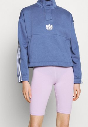 Person trägt blauen, langärmeligen, bauchfreien Hoodie mit weißem Logo und Streifen an den Ärmeln, kombiniert mit hellvioletten Leggings bis zum Knie.