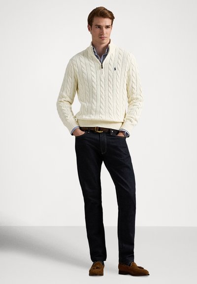 Kremfarget kabelstrikket genser med halv-zip-krage, kombinert med mørke jeans og brune mokkasin-sko, med en blå og hvit rutete skjorte under.