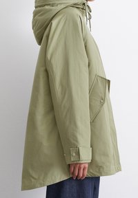 Khaki huva parka jacka med en avslappnad passform, med dragsnören i halsen, stora fickor med lappar och tryckknappsmuddar. Slät textur.