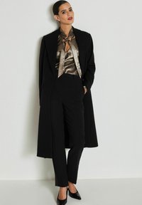 Schwarzer maßgeschneiderter Mantel, metallisch bronzefarbene Bluse mit Schlitzdetails und schwarze maßgeschneiderte Hose. Glänzende schwarze High Heels runden das Outfit ab.