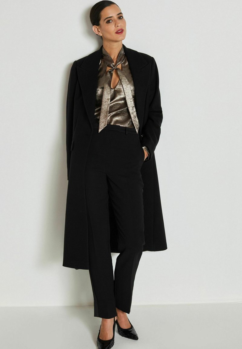 Schwarzer maßgeschneiderter Mantel, metallisch bronzefarbene Bluse mit Schlitzdetails und schwarze maßgeschneiderte Hose. Glänzende schwarze High Heels runden das Outfit ab.