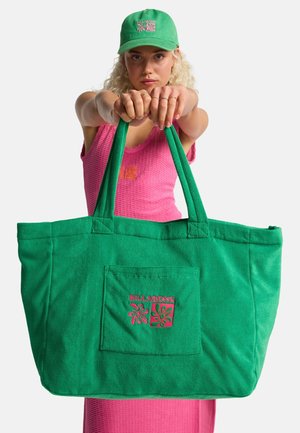 Blonde vrouw in een roze jurk en een groene pet die een grote groene Billabong draagtas met voorvak en roze logo vasthoudt.