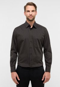 Camicia nera con bottoni, a righe verticali strette in arancione e beige. Presenta un colletto standard e una tasca sul petto. Texture liscia.