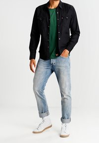 Camisa negra de manga larga superpuesta sobre una camiseta verde, combinada con jeans azul claro enrollados y zapatillas blancas con suelas marrones.