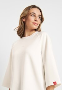 Ecru sweatshirt met een ronde hals, raglanmouwen en een klein roze logo. Heeft een rode accentlabel op de linkermouw. Soepele textuur.