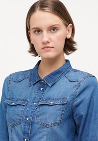 Πουκάμισο από τζιν με κουμπιά, διαθέτει δύο θήκες στο στήθος, κομψό γιακά και διακριτές λεπτομέρειες ραψίματος. Το ύφασμα είναι μέτριου μπλε χρώματος με λεία υφή.