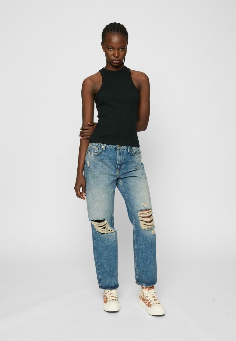 Svart ärmlös topp, ljusblå slitna jeans med stora hål och vita sneakers med bruna accenter. Enkel, figurnära design.