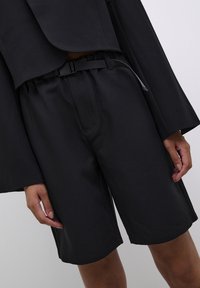 Pantalones cortos negros de tela suave, con un diseño frontal plano, una cinturilla elástica con un cinturón y bolsillos laterales.