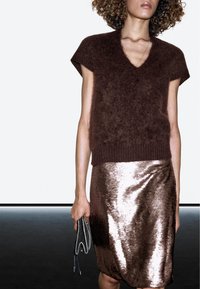 Brauner, flauschiger Pullover mit kurzen Ärmeln und V-Ausschnitt, kombiniert mit einem glänzenden Bleistiftrock aus rosegoldenen Pailletten. Hält eine kleine, graue Clutch in der Hand.