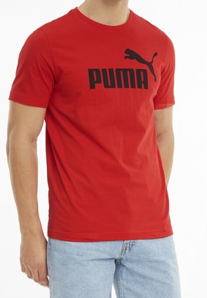 Homme portant un t-shirt rouge Puma avec un logo noir et un jean bleu clair, torse et parties supérieures des jambes visibles.