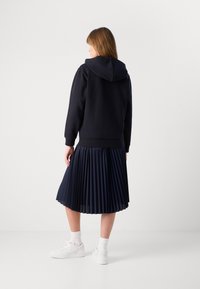 Sudadera con capucha de color navy combinada con una falda navy con pliegues. La modelo lleva zapatillas blancas y calcetines hasta el tobillo. Tejido suave con pliegues estructurados.