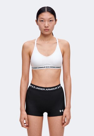 Woman standing facing forward wearing a white Under Armour sports bra and black Under Armour shorts against a plain background.