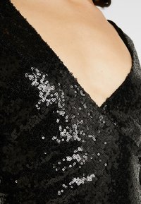 Svart paljettklänning med djup V-ringning och figurnära design. Texturen är glänsande med reflekterande paljetter som är tätt arrangerade.