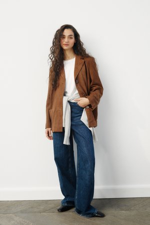 Brauner Wildlederblazer mit zwei Knöpfen, kombiniert mit einem weißen T-Shirt, einem grauen Pullover, der um die Taille gebunden ist, und weit geschnittenen blauen Jeans. Schwarze Schuhe.