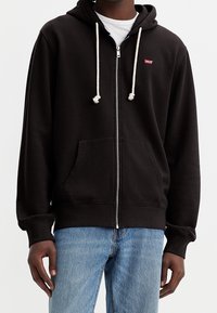 Zwarte hoodie met rits, gemaakt van katoen, met een capuchon, witte koordjes, een zak aan de voorkant en een klein rood logo op de borst.