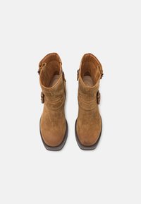 Brown suede ankle boots feature a rounded toe, side zip, and a decorative strap with a buckle. The sole is dark and slightly elevated.
