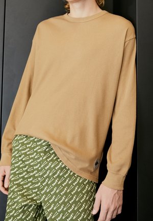Personne portant une chemise à manches longues beige et un pantalon vert avec le texte blanc "huf" et des motifs de clés à molette, debout devant un fond sombre.