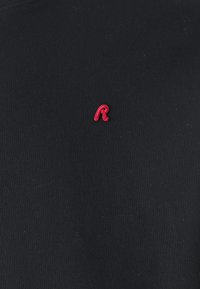 Sudadera negra con una letra 'R' bordada en rojo en el lado izquierdo del pecho. Textura suave, diseño de cuello redondo y mangas largas.