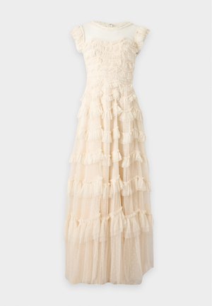 Femme debout portant une robe crème sans manches longue jusqu'au sol avec des volants superposés et un tissu transparent à pois, associée à des talons pointus beige.