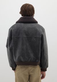 Giubbotto bomber in pelle nera con colletto in shearling marrone, polsini e vita a coste. Texture liscia, dettaglio della toppa sulla spalla visibile.