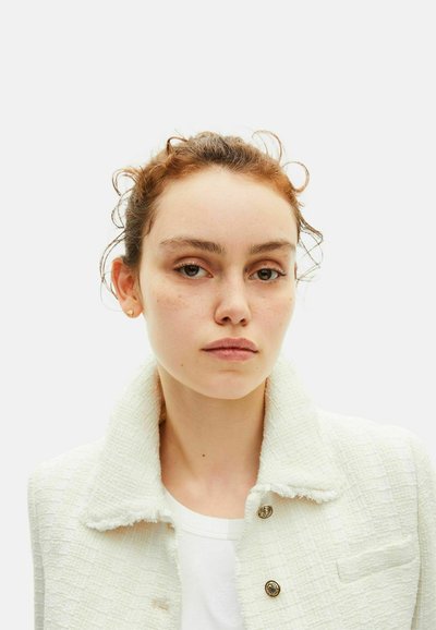 Jeune femme à la peau claire et aux cheveux roux, portant une veste blanche texturée avec des boutons dorés, sur un fond clair uni.
