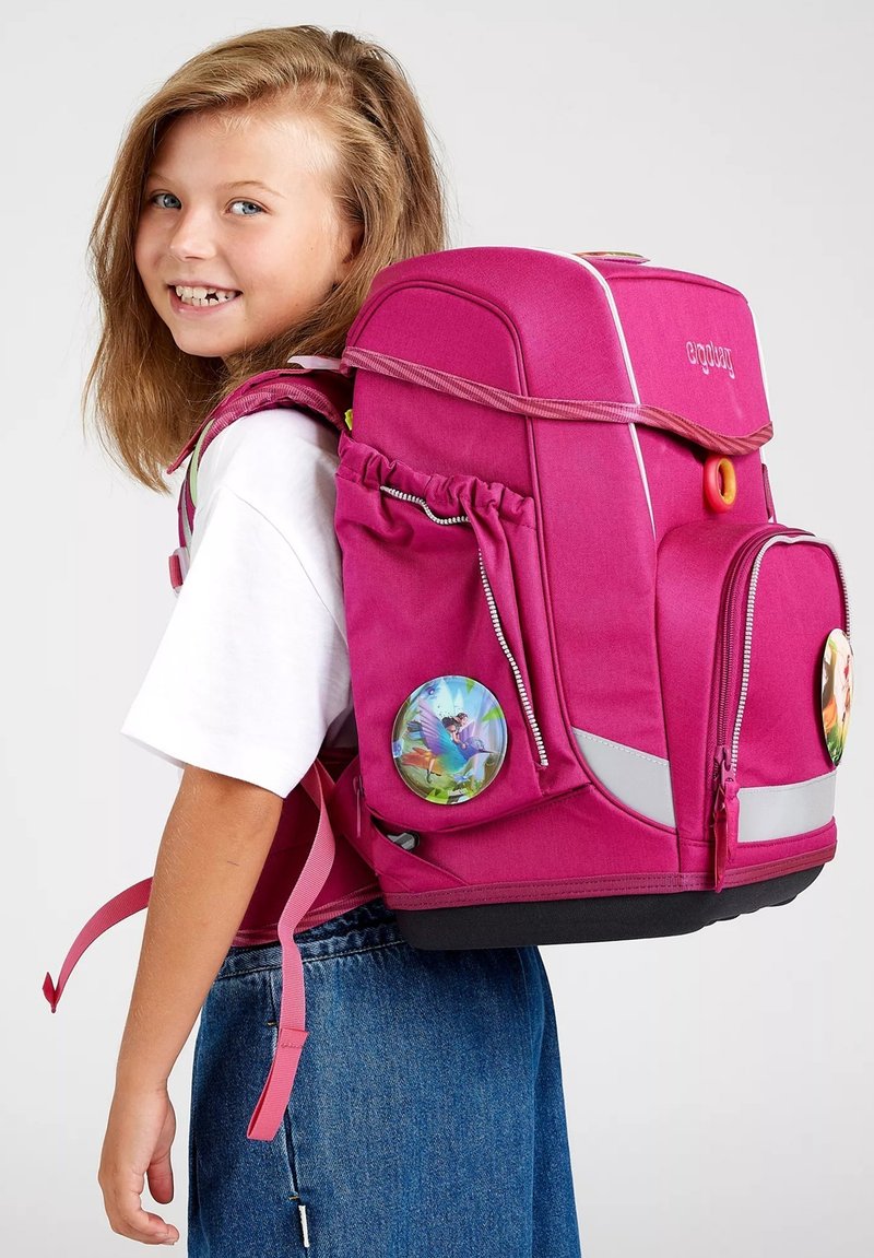 Zaino fucsia con molte tasche, strisce riflettenti e grandi toppe rotonde con design colorati, indossato da una ragazza in una maglietta bianca.