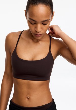 Femme en forme portant un soutien-gorge de sport marron, regardant vers le bas, une main touchant son cou sur fond blanc.