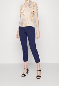 Blommig blus med volangärmar, ljus persikofärgad bas; marinblå capribyxor; svarta sandaler med remmar; slät textur; elegant rosettdetalj vid halslinjen.