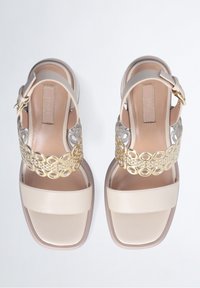 Beige sandalen met een vierkante teen, voorzien van een gouden cirkelpatroon over de bovenste band en transparante accenten aan de zijkanten. Rubberen zool.