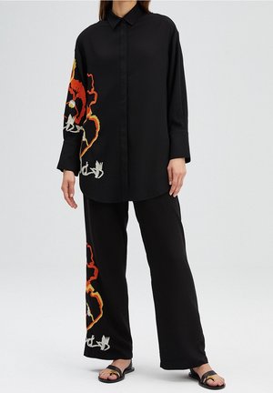 Mujer vestida con camisa y pantalones negros y holgados con un gran bordado floral naranja, amarillo y blanco en la manga izquierda y la pierna del pantalón, y sandalias negras.