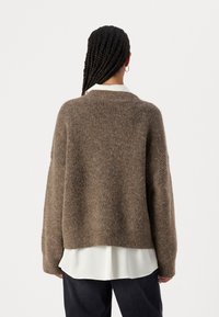 Brun strikket genser med avslappet passform, rund hals og lange ermer, lagdelt over en hvit bluse og parret med mørke jeans.