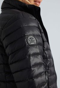 Gros plan sur une personne portant une veste matelassée noire avec un patch rond du logo montagne sur le haut du bras et une fermeture éclair à l'avant.