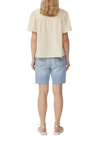Top corto a maniche corte di un beige chiaro e tessurizzato, con una vestibilità ampia, abbinato a pantaloni shorts in denim blu chiaro con orli sfilacciati e sandali bianchi.