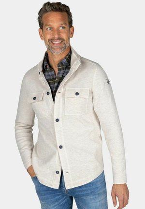 Smiling man wearing a light beige button-up jacket over a dark plaid shirt and blue jeans, standing with one hand in his pocket.