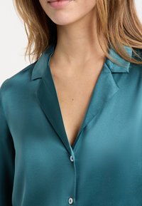 Blusa de satén en color teal con cuello, que presenta un escote en V y cierre de botones, mostrando una textura lisa y brillante.