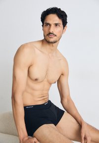 Uomo senza maglietta con capelli scuri e barba curata, indossa boxer neri GANT, seduto su uno sfondo chiaro e uniforme.