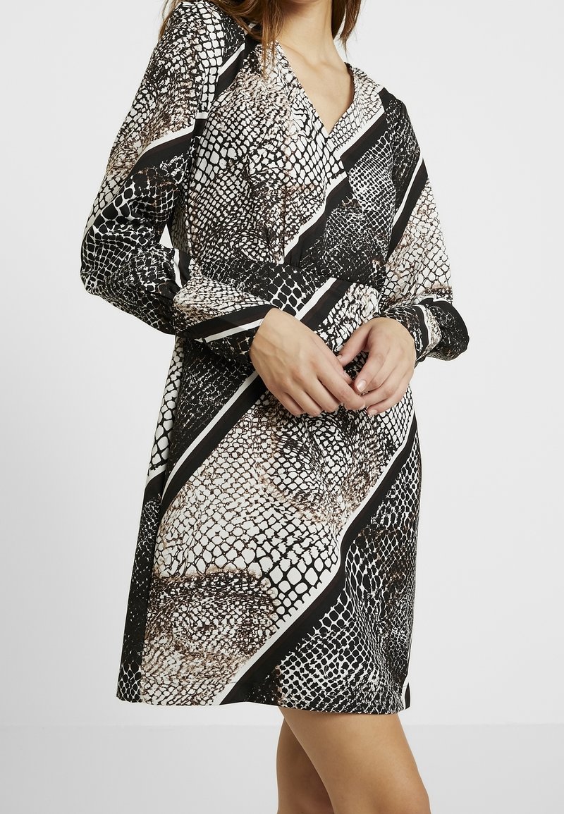 Femme portant une robe longue à manches longues imprimée serpent noir et blanc avec un col en V, debout, mains jointes près de la taille.
