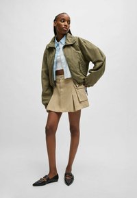 Oliven grøn bomberjakke, lyseblå rutet crop-top, beige plisseret nederdel med lomme og sorte spidse flade sko. Glatte teksturer gennemgående.