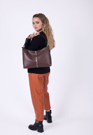 Femme aux longs cheveux blonds portant un pull noir, un pantalon orange, des bottes noires, tenant un grand sac à main en cuir marron sur son épaule.