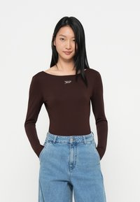 Bodysuit marrón de manga larga con escote en forma de barco y un pequeño logo plateado en el pecho, combinado con unos vaqueros de denim azul claro de cintura alta.